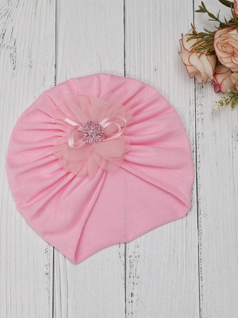 A light pink turban for baby girls featuring a soft cotton fabric and a decorative flower, presented on a rustic wooden background