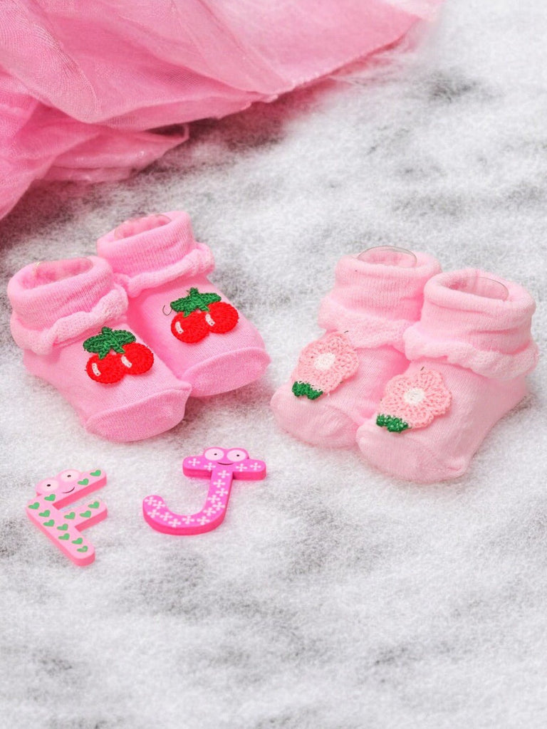 Pink baby socks with cherry and flower appliqués on a sparkly background with playful toys.