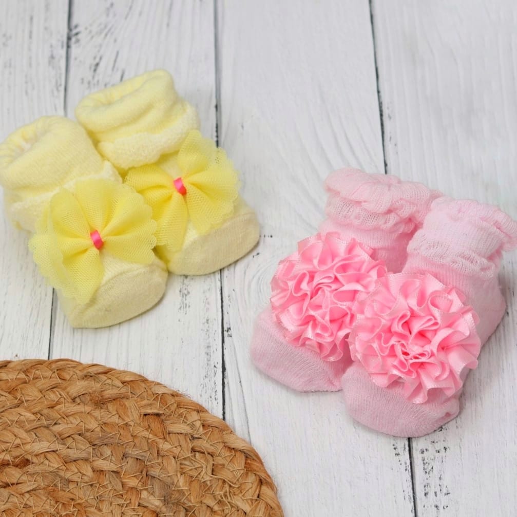 Two pairs of baby girl socks with ruffled flower details in yellow and pink on a rustic white wood background.