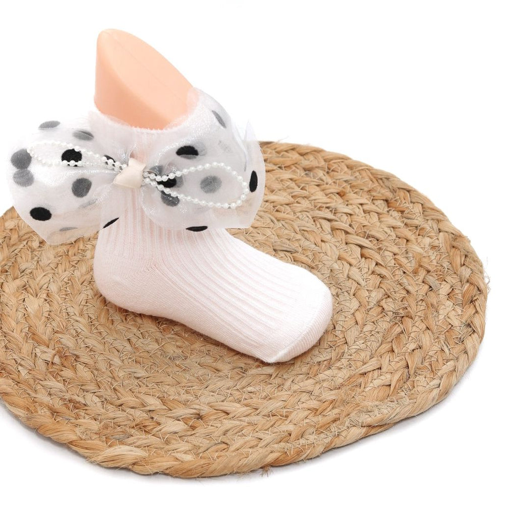 Child's white sock with polka dot bow and pearl detail on mannequin foot against a straw mat background.