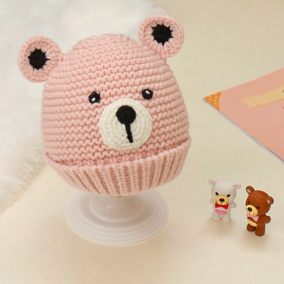 Handcrafted pink crochet teddy bear beanie for baby girls with adorable ear accents on a display stand.