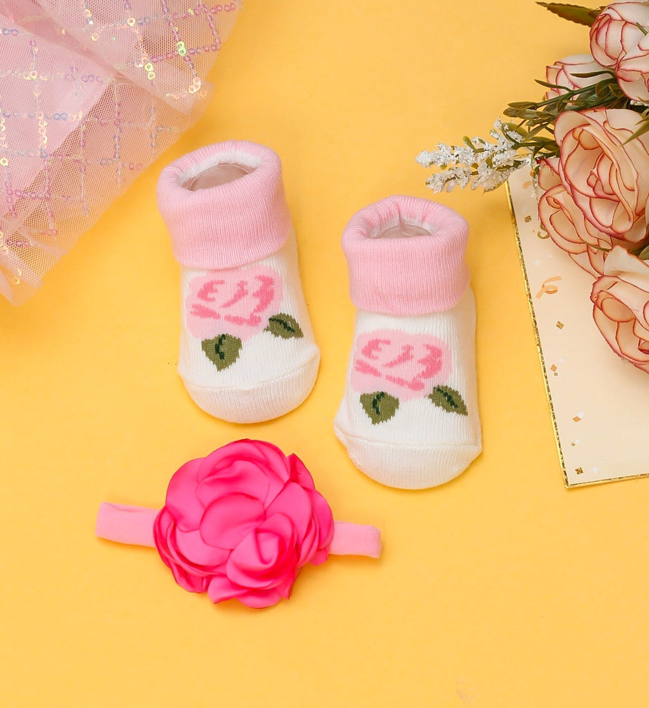Pink and white baby socks with rose patterns, paired with a vibrant pink flower headband, set against a yellow background.