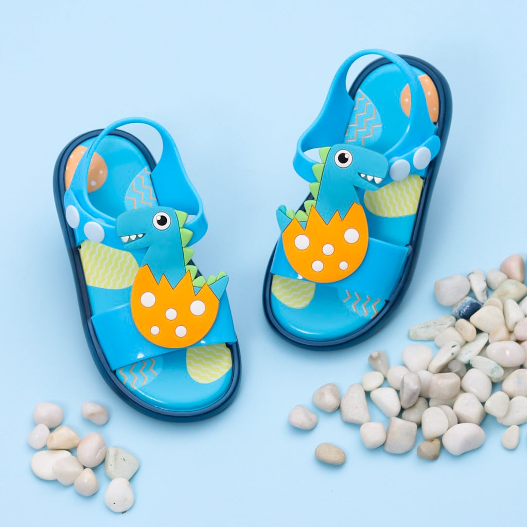 Blue dino-themed sandals for toddlers with playful design on a background of pebbles.