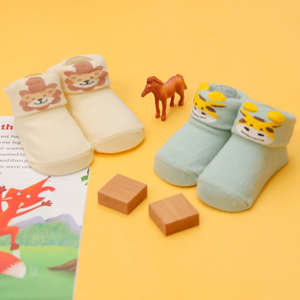 A pair of cream-colored infant socks with lion print and a pair of light blue socks with giraffe print against a yellow background.