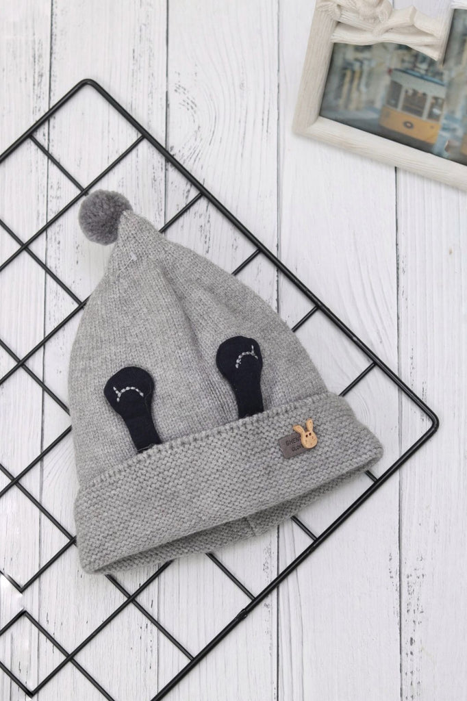 Grey winter beanie with eye design and pom-pom on a white wooden background.