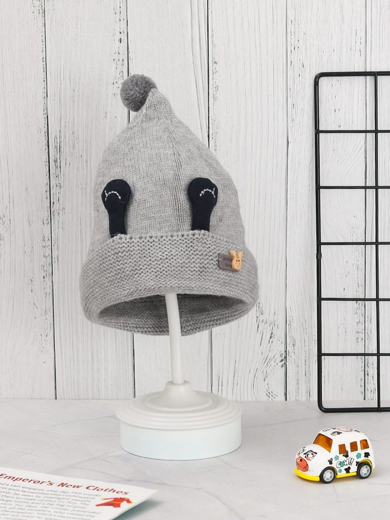 Grey winter beanie with eye appliques and pom-pom for boys on display stand.