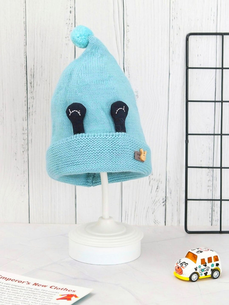 Light blue winter beanie hat with eye applique and pom-pom for boys displayed on a stand.