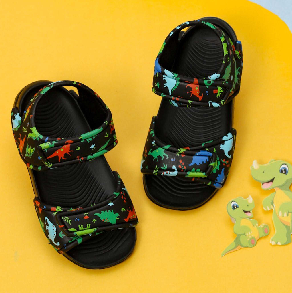 Pair of black sandals for kids with a colorful all-over dinosaur print on a yellow background