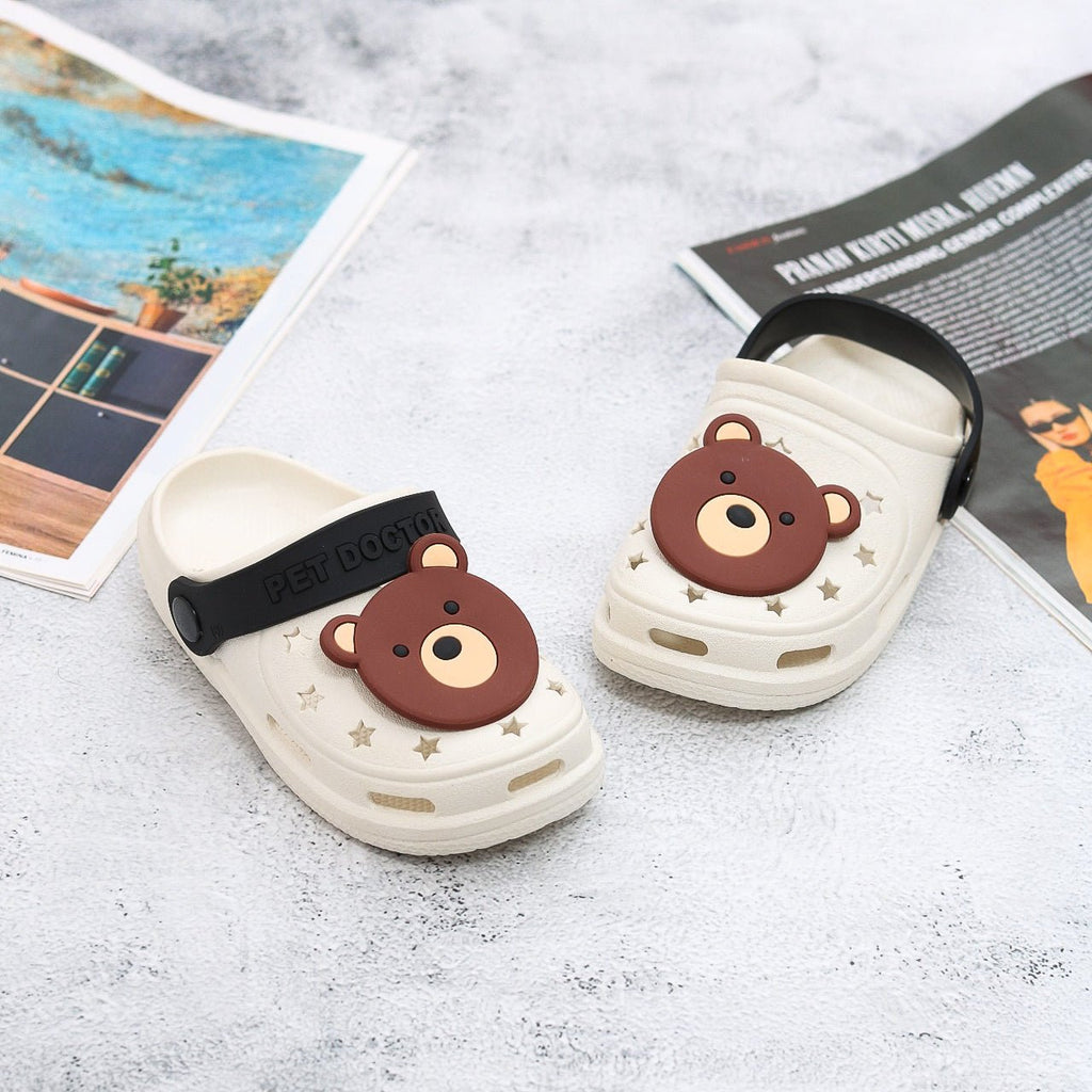 Pair of beige clogs with bear motif and black strap for children on a white background.