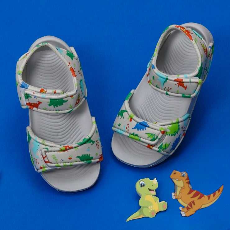 Children's grey sandals with a lively all-over dinosaur print, presented on a bold blue background.