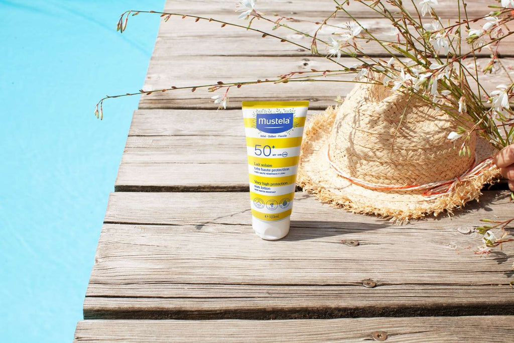 Mustela Very High Protection Sun Lotion SPF50+ next to a straw hat on a sunny poolside wooden deck