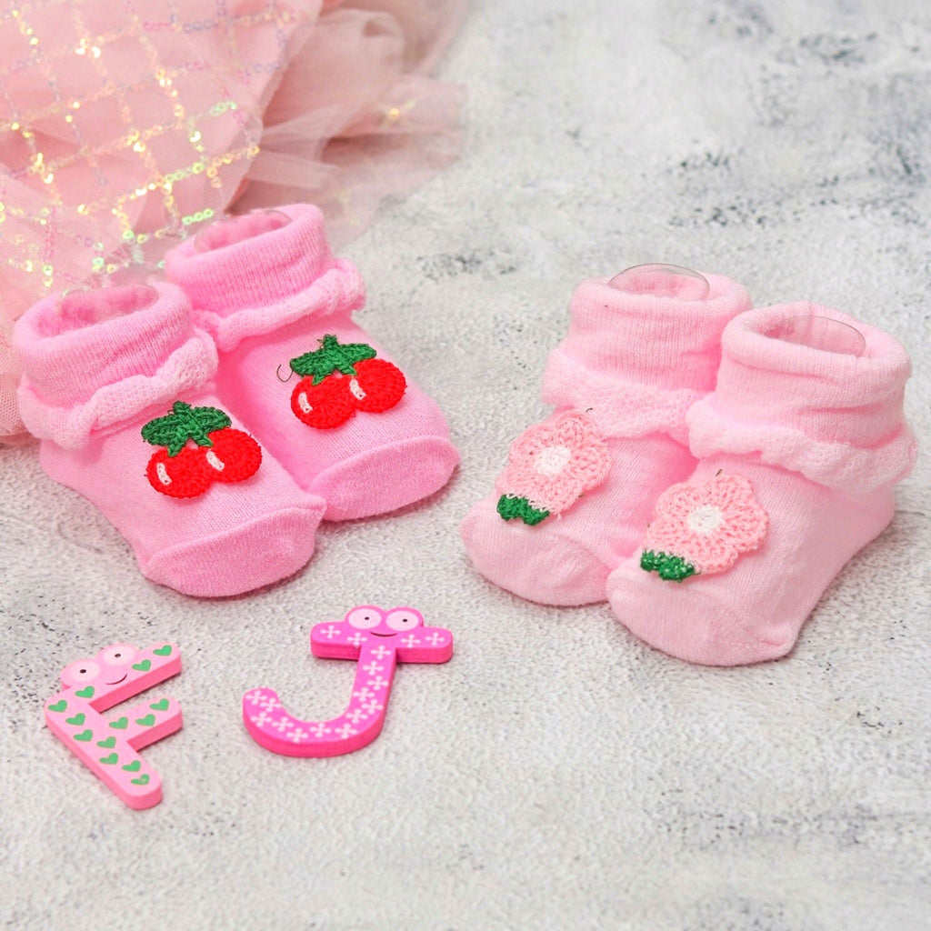 Pink baby socks with cherry and flower appliqués on a sparkly background with playful toys.