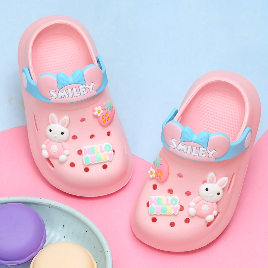 Pair of pink bunny motif clogs for kids with blue 'SMILEY' strap on pastel background.