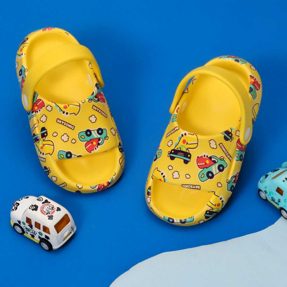 Bright yellow children's sandals with a playful dinosaur driving cars print against a vibrant blue background.