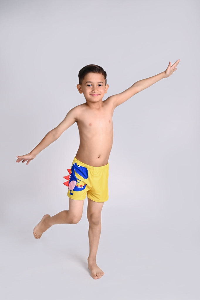 Young boy joyfully posing in yellow dinosaur-themed swim shorts with a blue and red side fin design