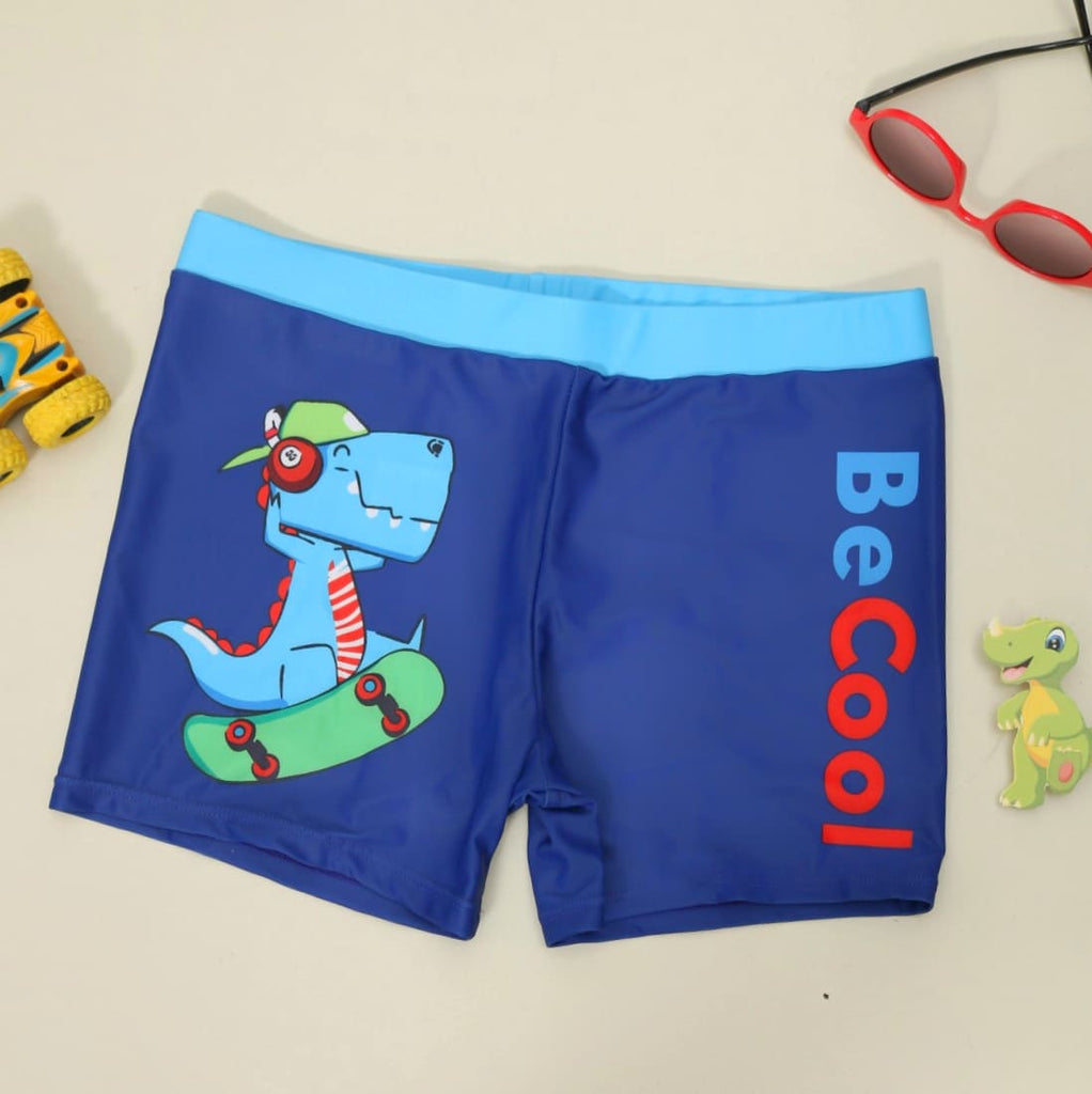 Blue swim shorts for boys with a playful skating dinosaur print and 'Be Cool' slogan, laid out flat 